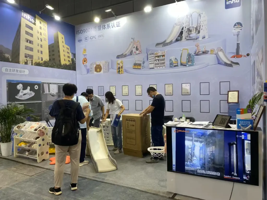 A busy view of the Inhe booth at CBME showing children's indoor slides, organizer racks, a boxed package, and a digital monitor playing factory manufacturing videos.