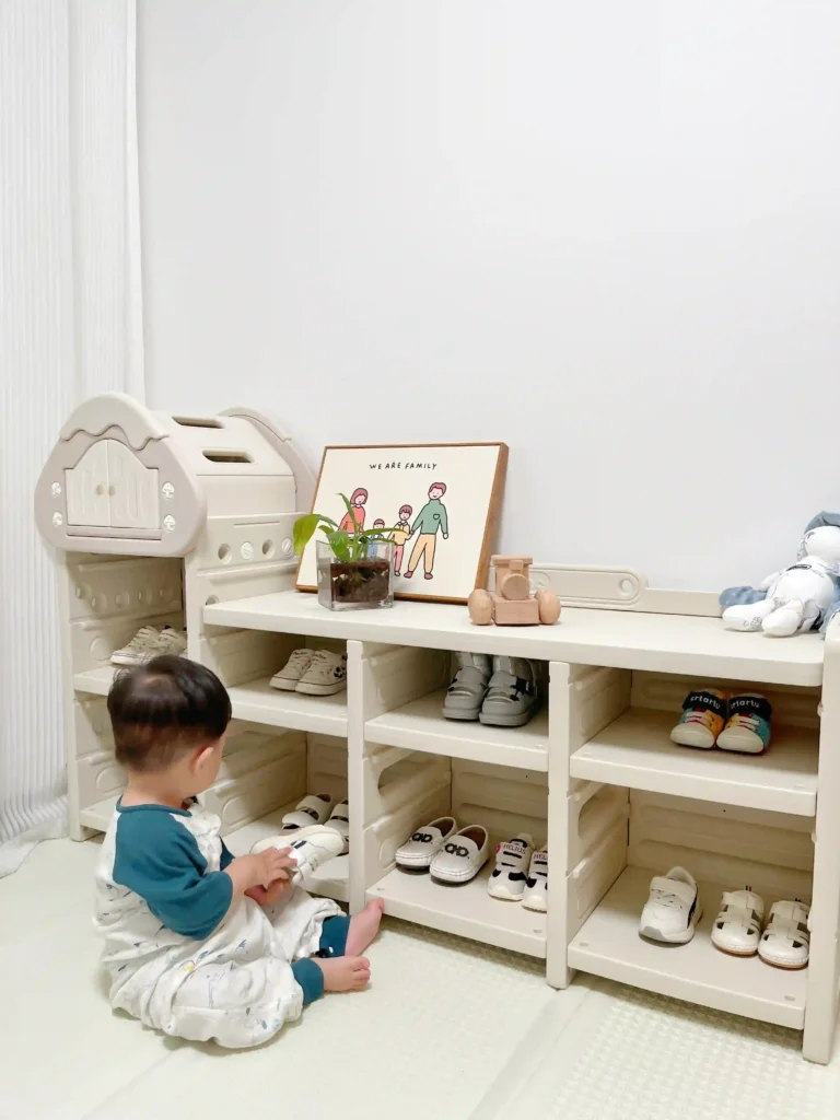 A toddler sitting on a play mat next to a minimalist, cream-colored children's storage cabinet and multi-tier shoe rack filled with toddler shoes.