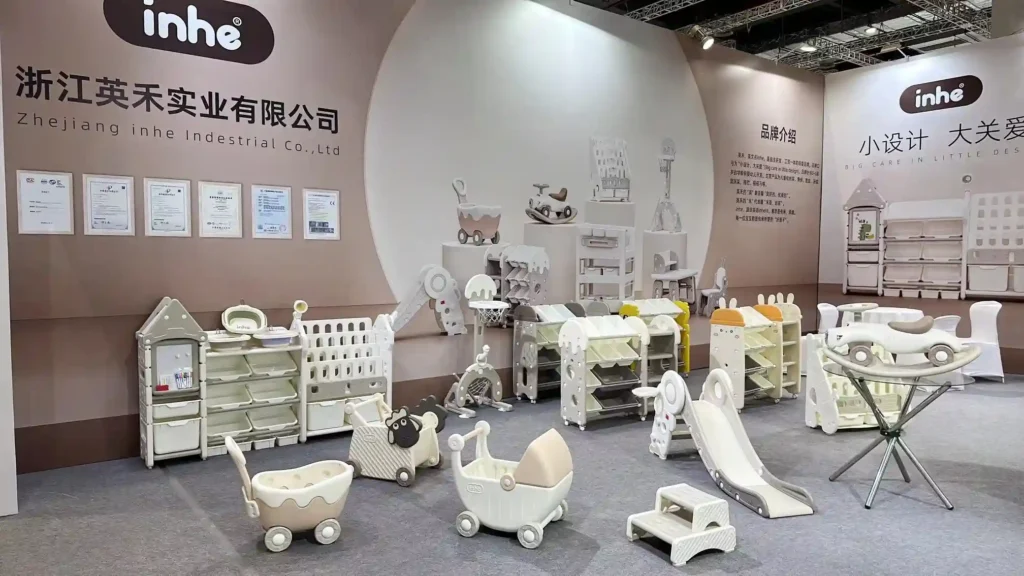 An interior view of the Inhe booth at CBME showing an organized arrangement of minimalist children's slide sets, feeding cabinets, house-shaped organizers, toy strollers, and small basketball hoops in soft cream tones.